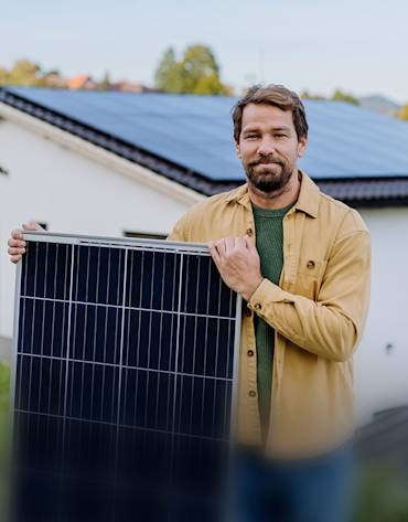 Man in a tan jacket holding a solar panel in front of a house with panels on the roof