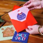 Handen die een zelf ontworpen kerstkaart in een rode envelop steken, met andere kerstkaarten op tafel
