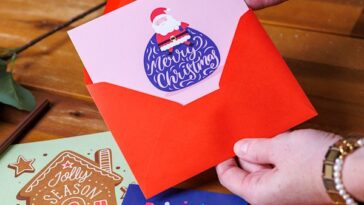 Handen die een zelf ontworpen kerstkaart in een rode envelop steken, met andere kerstkaarten op tafel