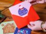 Handen die een zelf ontworpen kerstkaart in een rode envelop steken, met andere kerstkaarten op tafel