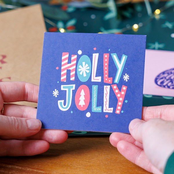 Vierkante kerstkaart met Holly Jolly tekst en een gekleurde kern in de hand